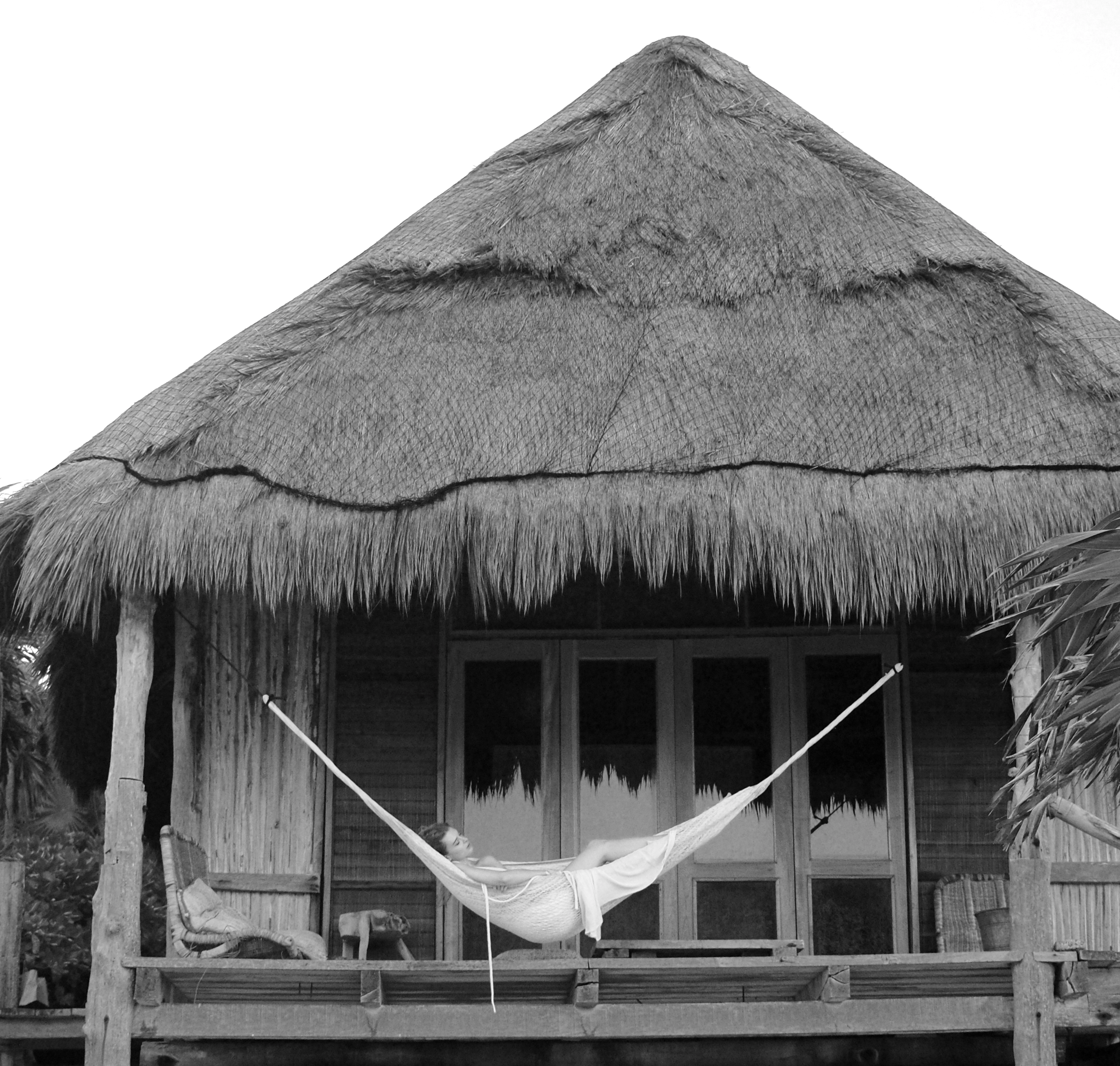 A woman relaxes in a hammock on the porch of a rustic hut, surrounded by greenery and sunlight.