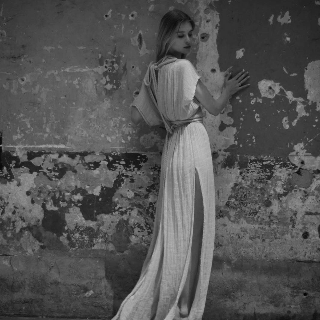 Lara Wegener in a long, light-colored dress standing against a textured wall.