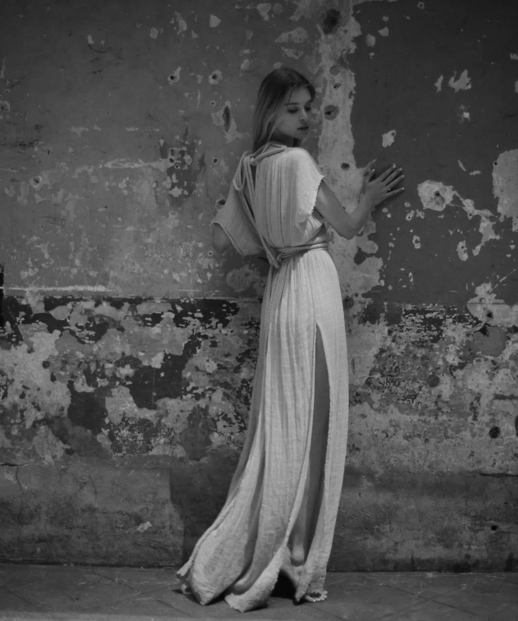 Lara Wegener in a long, light-colored dress standing against a textured wall.